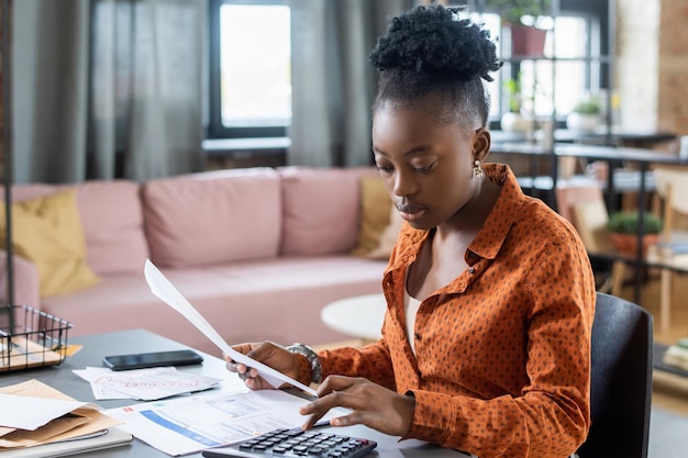 Femme noire concentrée en chemise décontractée assise à table dans le salon et calculant la facture d'impôt