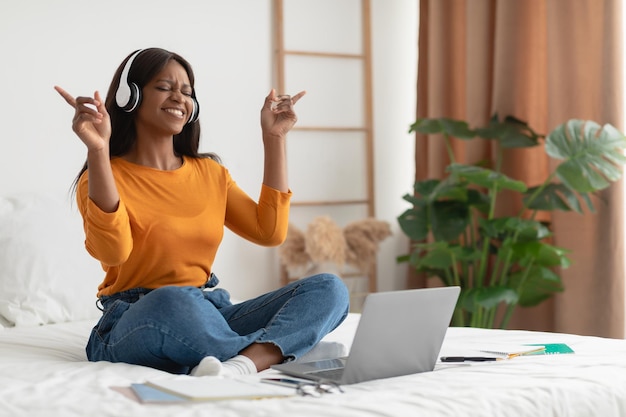 Femme noire écoutant de la musique à l'aide d'un ordinateur portable et d'un casque dans la chambre