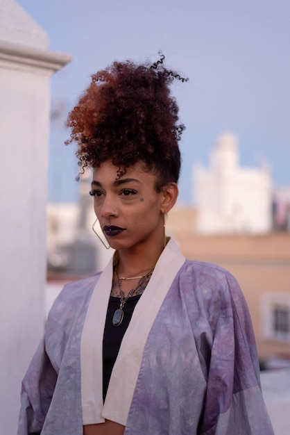 Femme noire élégante aux cheveux bouclés et aux lèvres noires portant un kimono tout en se reposant sur une terrasse contre un ciel bleu sans nuages à Cadix en Espagne