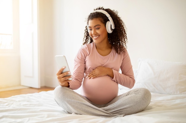 Une femme noire enceinte heureuse apprécie la musique en utilisant un smartphone et des écouteurs sans fil