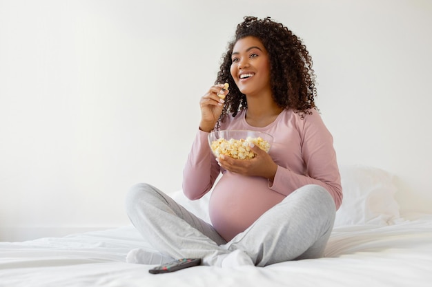 Une femme noire enceinte joyeuse mangeant du pop-corn assise sur le lit à la maison.