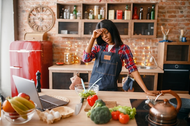 Femme noire endormie en tablier, cuisine petit-déjeuner sain dans la cuisine. Femme africaine préparant une salade de légumes à la maison