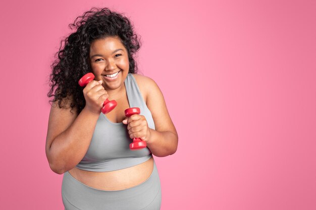 Une femme noire excitée en taille plus en vêtements de sport tenant des haltères et riant debout sur le rose