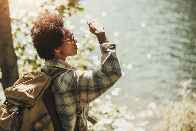 Femme noire mature avec sac à dos faisant une pause et profitant de la vue sur le lac lors d'une randonnée.