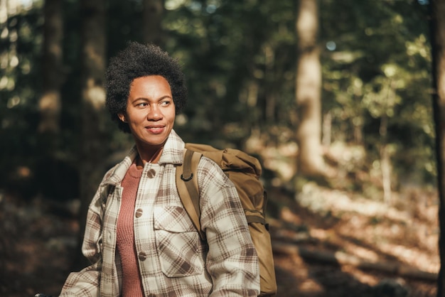 Femme noire mature avec sac à dos profitant de la vue sur la forêt lors d'une randonnée dans la nature.