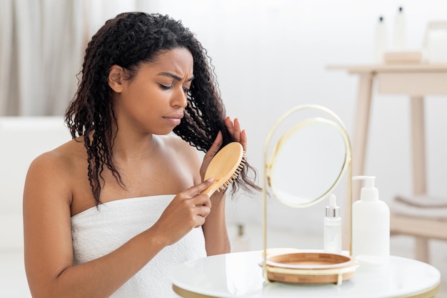 Femme noire mécontente peignant ses cheveux emmêlés avec une brosse en bambou