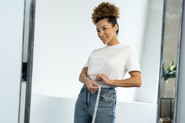 Une femme noire mince debout devant un miroir mesure sa taille avec un ruban à mesurer après