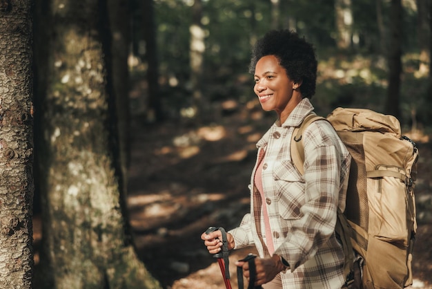 Femme noire mûre souriante, debout seule à l'extérieur et portant un sac à dos lors d'une randonnée dans les montagnes.