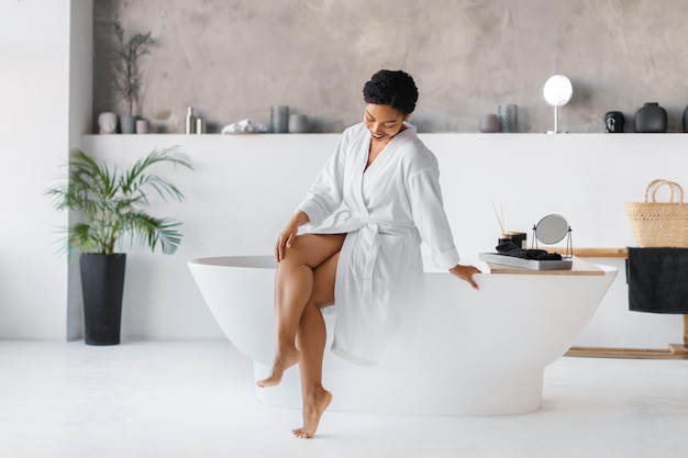 Une femme noire en peignoir assise sur la baignoire et touchant ses belles jambes.
