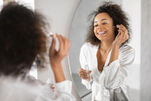 Une femme noire près du miroir applique un sérum cosmétique facial dans la salle de bain