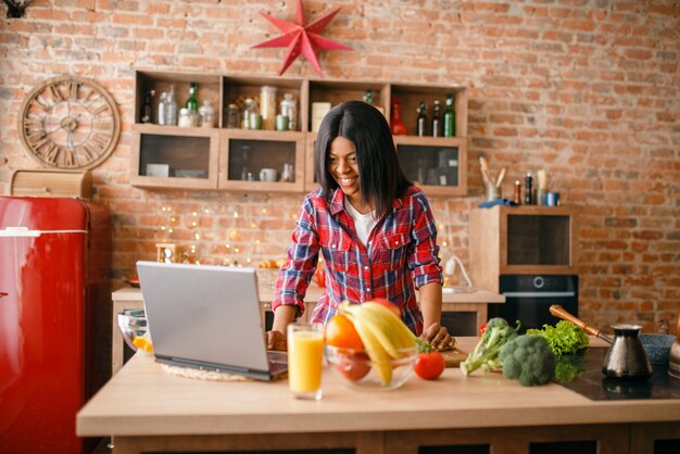 Femme noire à la recherche de recette sur ordinateur portable, jeune fille ethnique cuisine dans la cuisine