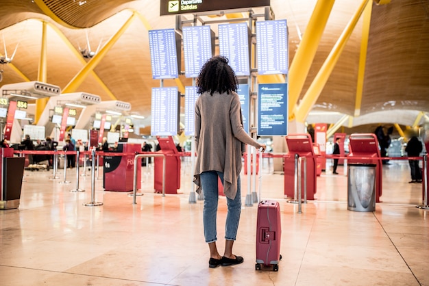 Femme noire en regardant le panneau d'information sur les horaires à l'aéroport avec une valise