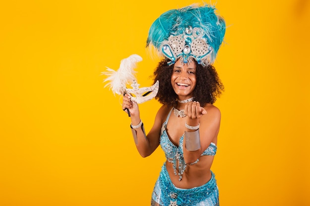 Femme noire reine de l'école de samba brésilienne avec des vêtements de carnaval bleus et une couronne de plumes tenant un masque invitant les mains