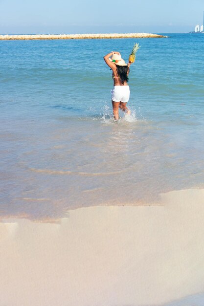 Une femme noire s'amuse sur la plage avec un ananas dans les mains.