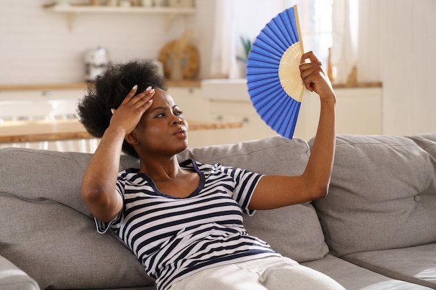 Femme noire souffrant d'un coup de chaleur à plat sans climatiseur agitant un ventilateur allongé sur un canapé à la maison