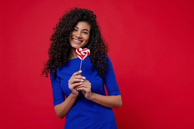 Femme noire souriante avec des bonbons de sucette en forme de coeur isolé sur mur rouge