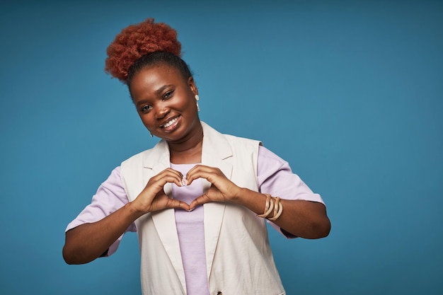 Femme noire souriante montrant le signe du coeur sur fond bleu en studio