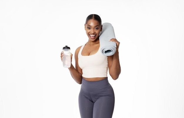 Une femme noire avec un tapis de fitness et une bouteille d'eau posant dans un studio