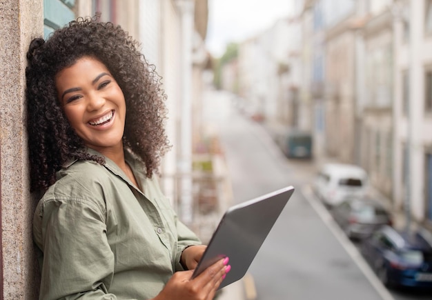 Femme noire tenant une tablette numérique debout sur le balcon de l'appartement