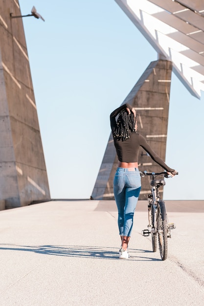 femme noire avec un vélo marchant et faisant rouler le vélo dans ses mains vue arrière