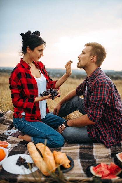 Photo femme nourrit des raisins à son homme en pique-nique dans le champ d'été. junket romantique de l'homme et de la femme, couple amoureux heureux ensemble