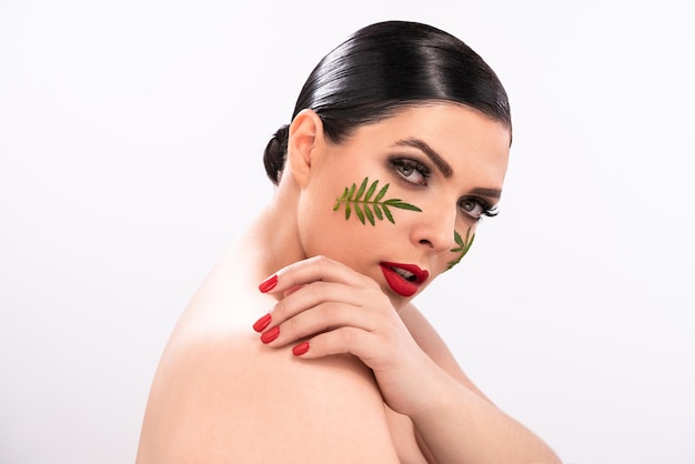 Femme nue sexy regardant la caméra par-dessus son épaule avec des taches de feuilles vertes sur son visage à base de plantes ca...