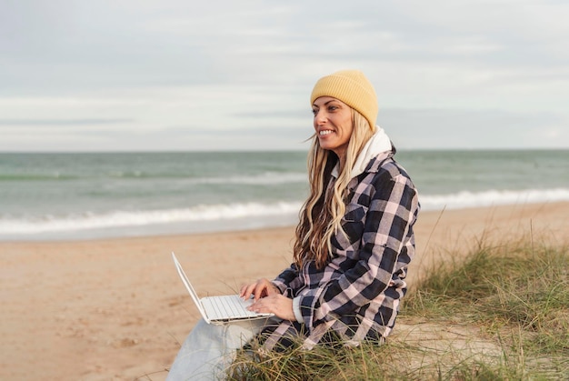 Femme numérique nomade voyageuse en hiver ou en automne assise sur la plage à l'aide d'un ordinateur portable