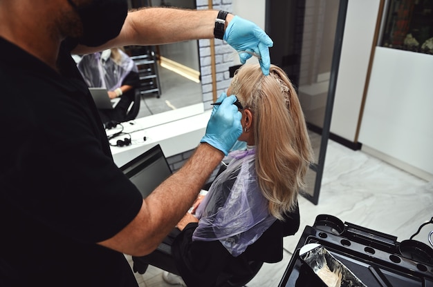 Femme occupée travaillant sur un ordinateur portable pendant que le coiffeur colorie une mèche de ses cheveux dans un salon de beauté