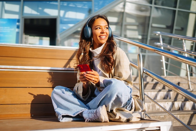 Femme optimiste portant des écouteurs et tenant le téléphone à l'extérieur Communication dans les réseaux sociaux