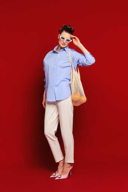 Femme avec des oranges dans un sac à cordes réutilisable sur l'épaule touchant ses lunettes de soleil