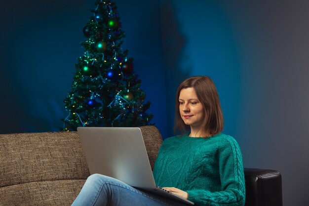 Femme avec un ordinateur assise sur un canapé et travaillant seule à la maison la veille de Noël