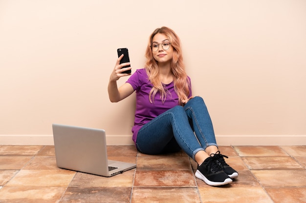 Femme avec un ordinateur portable assis sur le sol à l'intérieur faisant un selfie