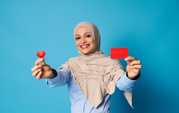 Femme orientale souriante en hijab montrant une forme de coeur rouge et carte en plastique rouge vierge, souriant