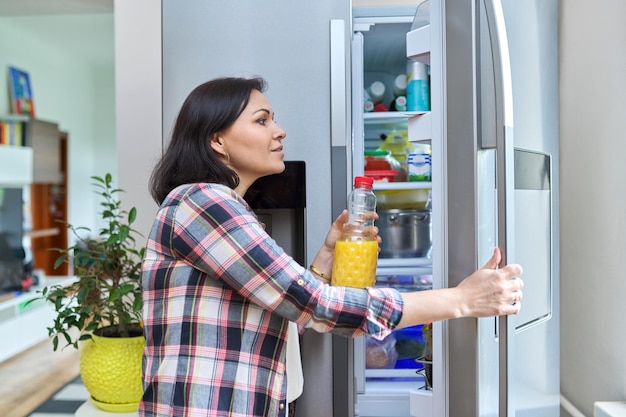 Une femme ouvre le réfrigérateur à la maison dans la cuisine tenant une bouteille de jus d'orange