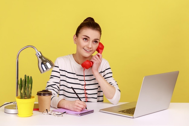 Femme Parlant Sur Un Téléphone Rétro Rouge A Une Expression Rêveuse Assise Sur Le Lieu De Travail Et écrivant Dans Le Bloc-notes