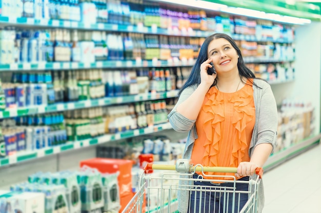 Femme parlant de téléphone et tenant un caddie dans le magasin de supermarché près des vitrines des magasins.