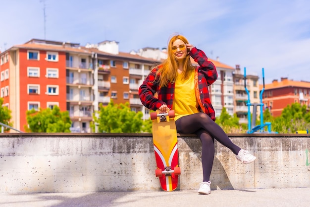 Femme patineuse dans un t-shirt jaune chemise à carreaux rouge et lunettes de soleil assis avec planche à roulettes sur un banc dans la ville souriant regardant la caméra