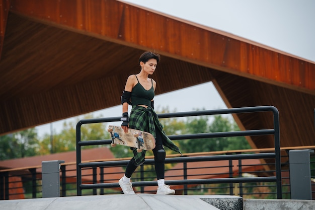 Femme patineuse descendant la rampe avec désinvolture avec sa planche à la main. Grand toit de forme créative derrière elle.