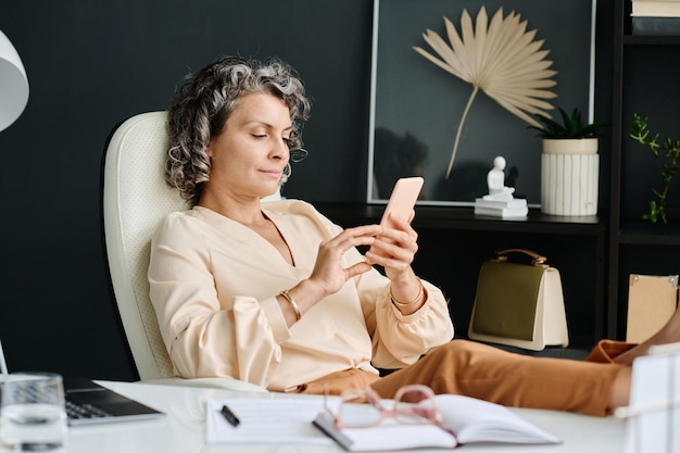 Femme PDG détendue et mature dans une tenue de luxe tranquille à l'aide de son téléphone portable