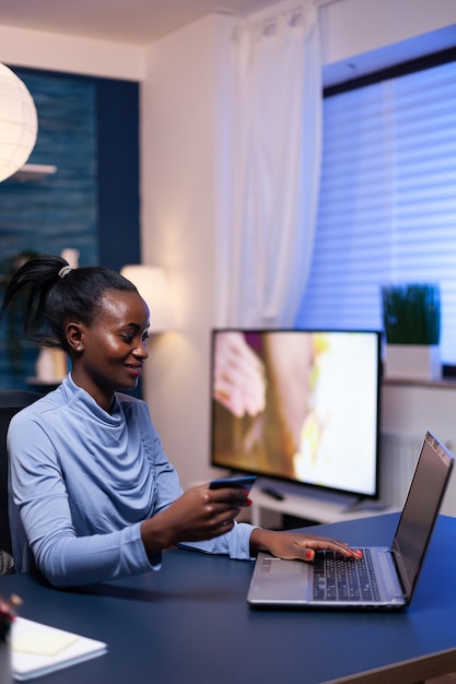 Femme à la peau foncée effectuant une transaction en ligne à l'aide d'une carte de crédit en plastique assise au bureau dans le bureau à domicile. Employé effectuant une transaction de paiement depuis son domicile sur un ordinateur portable numérique.