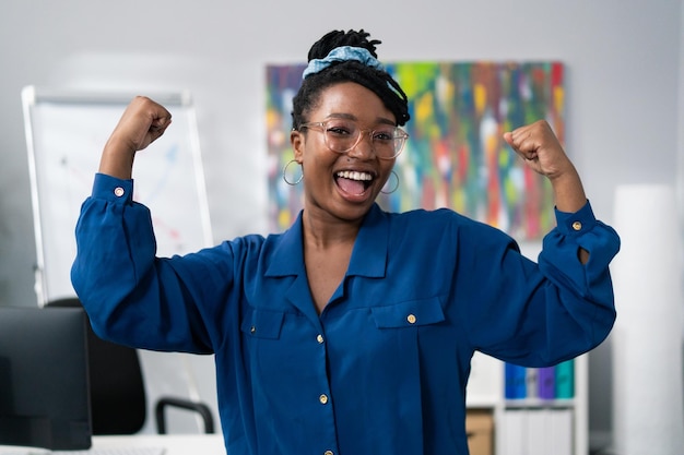 Une femme à la peau foncée portant des lunettes de vêtements bleus se dresse sur fond de bureau sourit heureuse victoire