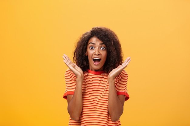 Femme à la peau sombre avec une coiffure afro en tshirt rayé avec des paumes levées près du visage à la caméra étonné