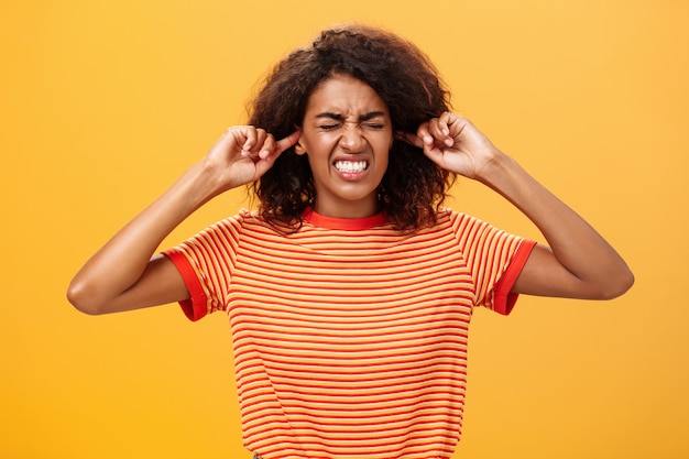 Photo femme à la peau sombre serrant les dents et fermant les oreilles agacé sur le mur orange