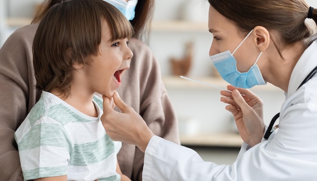 Femme pédiatre professionnelle portant un masque protecteur vérifiant la gorge d'un petit garçon patient