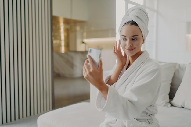 Une femme en peignoir applique de la crème touche une peau douce assise sur un canapé et tient la routine de beauté du smartphone