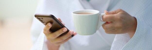 Photo femme en peignoir blanc buvant du café dans une tasse et tenant un téléphone portable à la maison agrandi