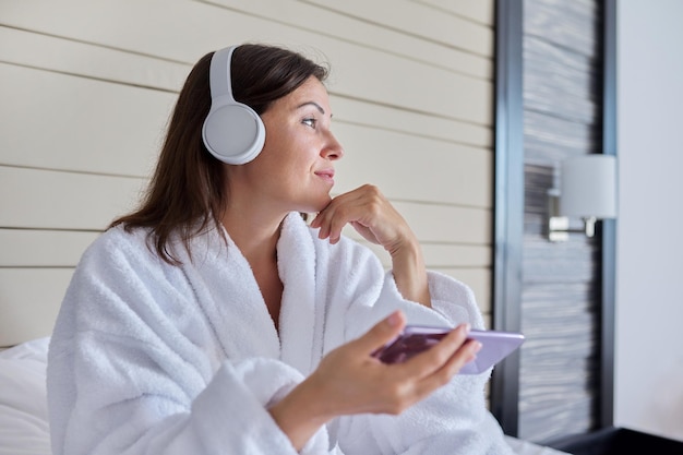 Femme en peignoir et écouteurs écoutant de la musique et de la détente assis dans son lit