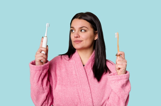 Une femme en peignoir rose tient à la fois une brosse à dents électrique et une brosse à dents traditionnelle