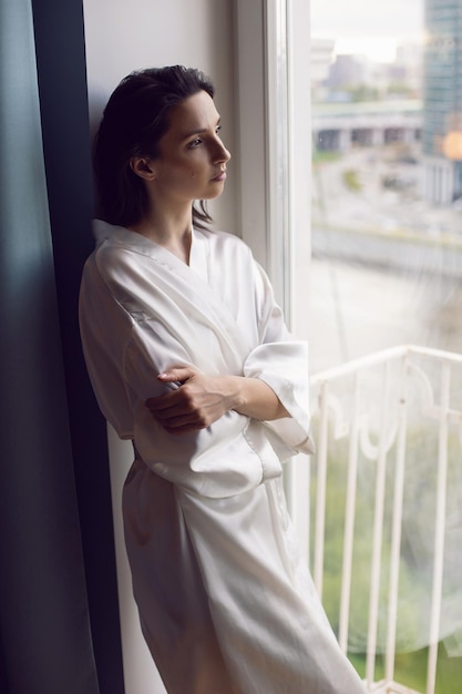 Une femme en peignoir se tient devant une grande fenêtre dans un appartement en face d'un gratte-ciel le matin