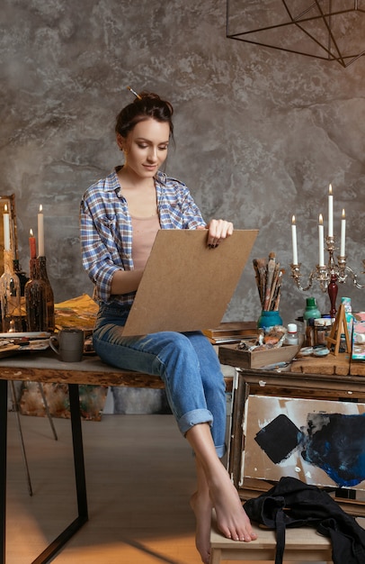 Femme peintre assise sur la table, dessinant et souriant. Créatif . Fournitures de dessin, peinture à l'huile, pinceaux d'artiste, toile, bougie, lampe à huile.